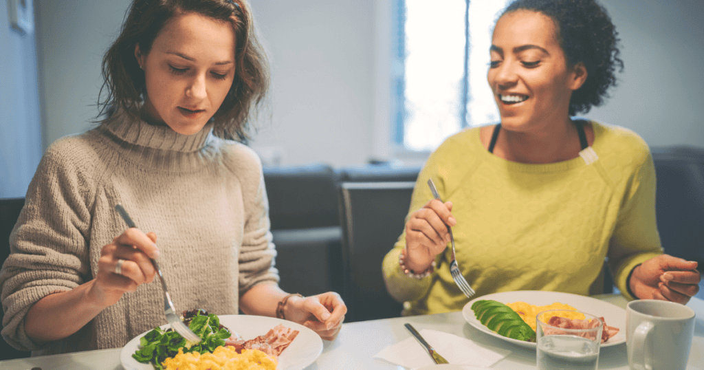 Two women sit at a table eating breakfast. One is wearing a beige sweater, the other a green top. Their plates have eggs, ham, and salad. One woman smiles while the other focuses on her food, offering subtle glimpses into possible warning signs of an eating disorder.