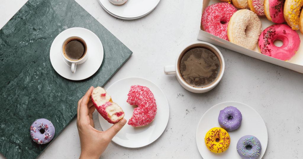 A hand holds a bitten pink-sprinkled donut above a plate; coffee cups, donuts on plates, and a donut box fill the table, creating a colorful breakfast scene that hints at an unhealthy relationship with food.