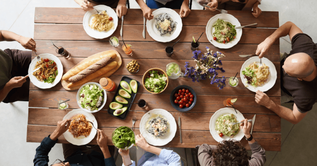 Eight people sit around a wooden table eating pasta and salad, demonstrating a healthy relationship with food. The table is set with bread, avocados, cherry tomatoes, flowers, and drinks. Plates are full, and everyone appears to be enjoying their meal together.