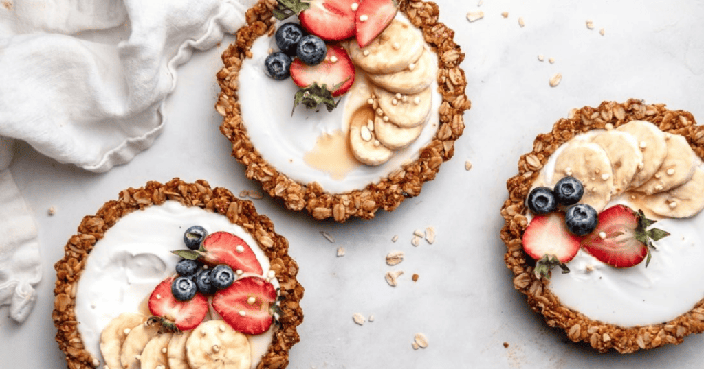Three granola tarts filled with yogurt, topped with banana slices, strawberries, blueberries, and a drizzle of honey are arranged on a white surface with scattered oats and a cloth napkin nearby—a nourishing treat that provides mindful enjoyment and can help you overcome binge eating by focusing on wholesome ingredients.