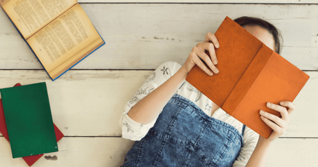 A person lying on a wooden floor, holding and reading an orange book that covers their face—perhaps one of those insightful books for emotional eaters. Other books, one open and two closed, are scattered nearby.