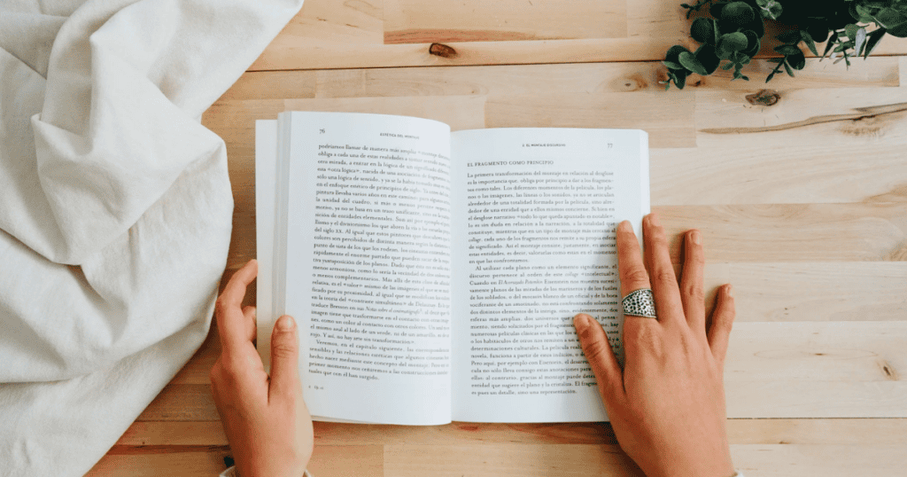 A person with a silver ring on one finger reads an open book on intuitive eating books at a light wooden table, with a white cloth and green plant nearby.
