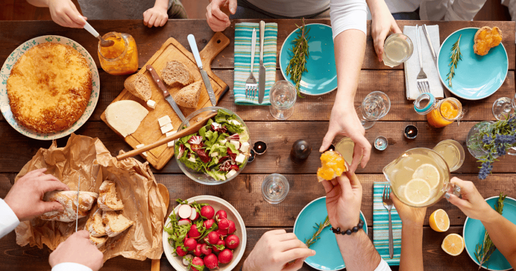 People share a meal at a rustic wooden table set with bread, salad, radishes, pie, drinks, plates, and utensils. Hands reach for food—some playfully acting as food pushers—creating a warm, communal dining atmosphere.