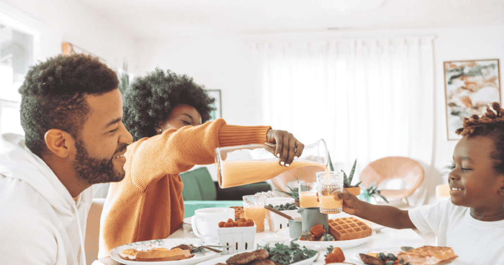A family of three sits at a breakfast table, smiling as they enjoy their morning together. One adult pours orange juice for the child, making navigating mealtime battles seem effortless. The table is filled with waffles, fruit, and other breakfast foods. The room is bright with natural light, creating a warm and inviting atmosphere.