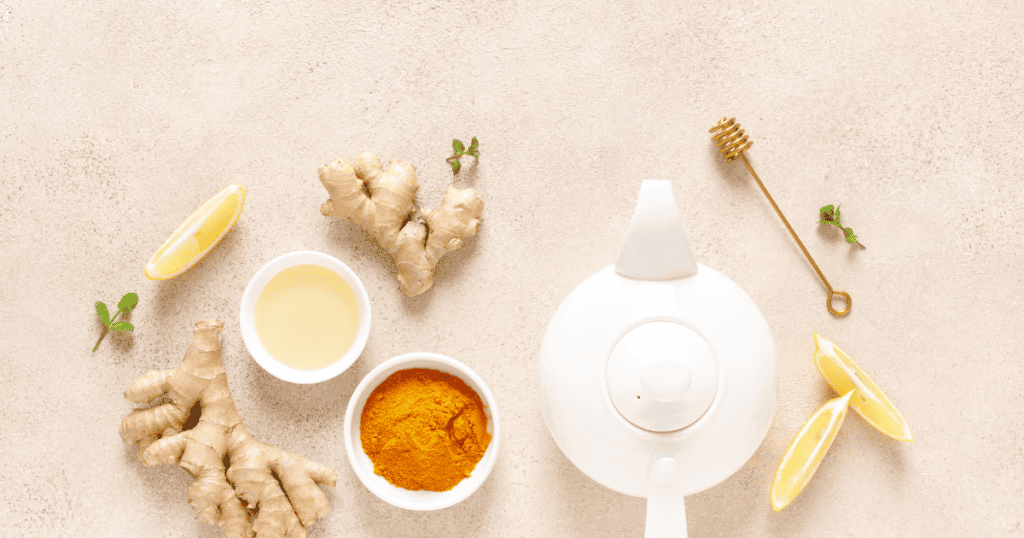 White teapot with ginger roots, lemon slices, a bowl of turmeric, a bowl of liquid, a honey dipper, and mint leaves arranged on a light beige surface—perfect elements for a natural detox ritual.