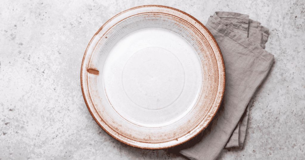 A large white ceramic plate with brown edging sits on a light gray textured surface, next to a neatly folded beige cloth napkin—perfect for illustrating topics like ARFID explained, where food presentation and mealtime environments play an important role.