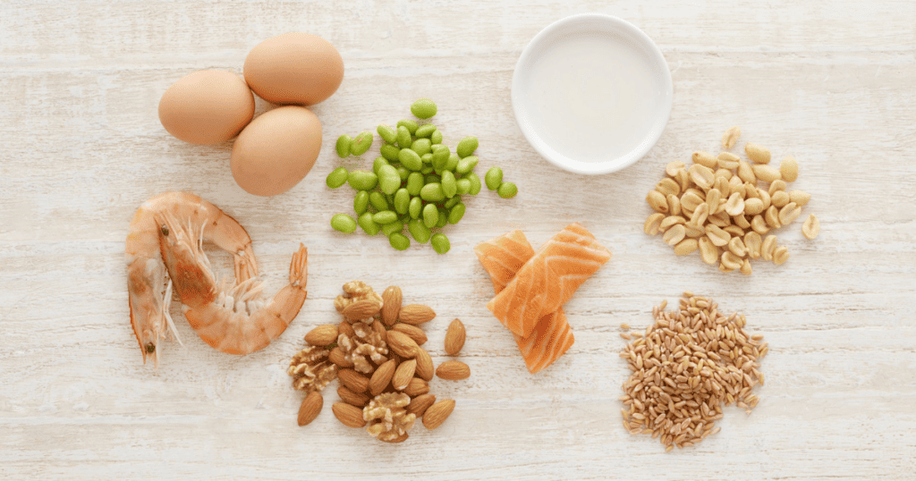 A selection of protein-rich foods on a white surface, including eggs, shrimp, edamame, nuts, salmon, seeds, and a small bowl of milk—each highlighting the important role of diet in managing PCOS by supporting balanced nutrition.
