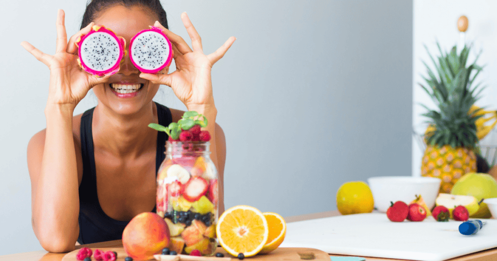 A smiling person holds two dragon fruit halves over their eyes, sitting at a table with various fresh fruits, a fruit jar, and a cutting board in a bright kitchen—a joyful celebration of the best nutrients for eye health and vision.