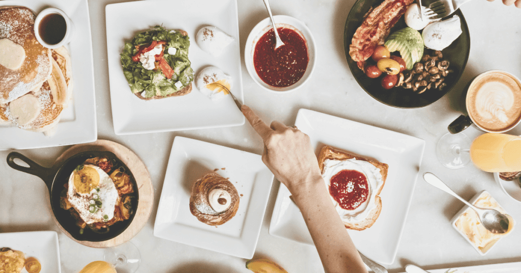 A table filled with various breakfast dishes, including toast, pancakes, eggs, fruit, coffee, and juice. Wondering how to stop the munchies? A hand reaches for food from a plate with tongs.