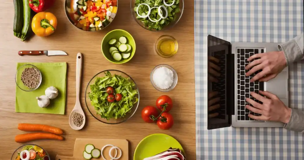 A person types on a laptop next to a table with fresh vegetables, bowls of salad, spices, and cooking ingredients suited for a FODMAP diet on a checkered tablecloth.