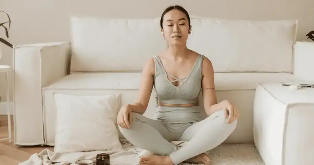A person in light athletic wear sits cross-legged on a rug, eyes closed, meditating in a calm living room with a white sofa and pillow in the background—cultivating peace and mindfulness to help manage migraine symptoms.