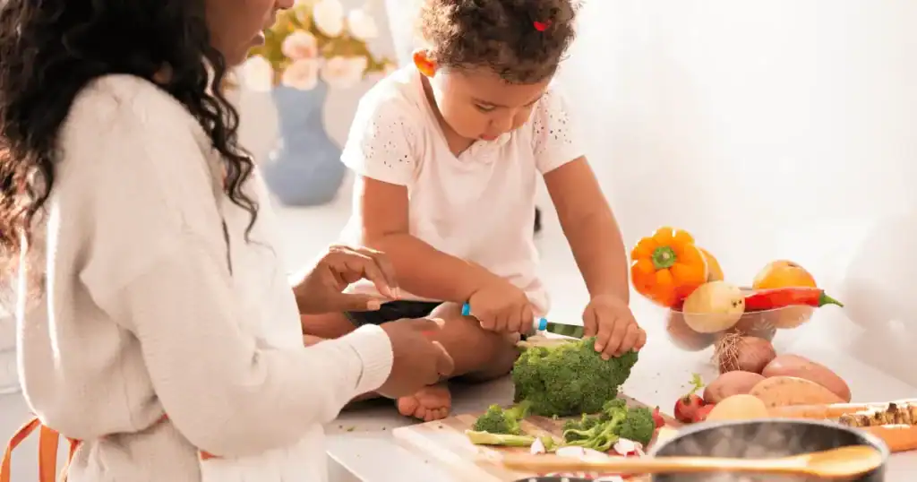 A young child sits on a kitchen counter cutting broccoli with help from an adult, taking the first steps toward raising healthy eaters. Various vegetables are spread on the counter around them.