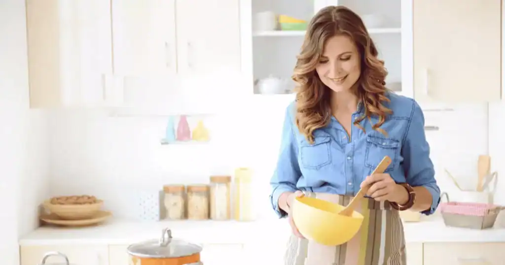A woman in a blue shirt and striped apron smiles while mixing ingredients in a yellow bowl in a bright, modern kitchen, thoughtfully preparing a meal that supports her nutritional needs during menopause.