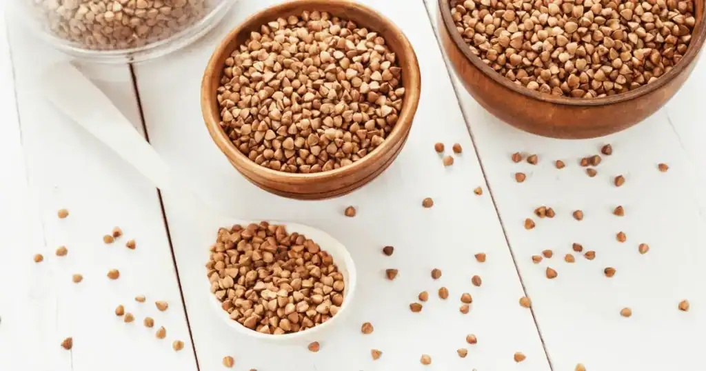 Several bowls and a spoon filled with dry buckwheat groats are arranged on a white wooden table, with some loose groats scattered around—an inviting scene for anyone interested in healthy eating and the benefits of a Glycemic Index Simplified approach.