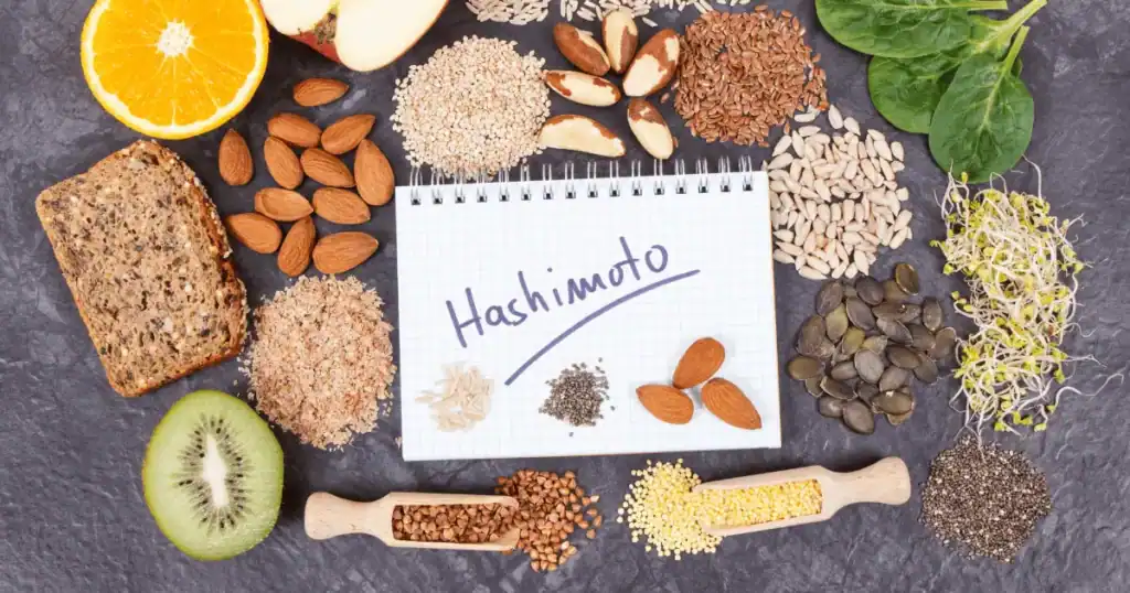 Various healthy foods like nuts, seeds, grains, fruits, vegetables, and whole grain bread surround a notepad with “Hashimoto” written on it on a dark surface, highlighting the importance of nutrition in supporting thyroid health.