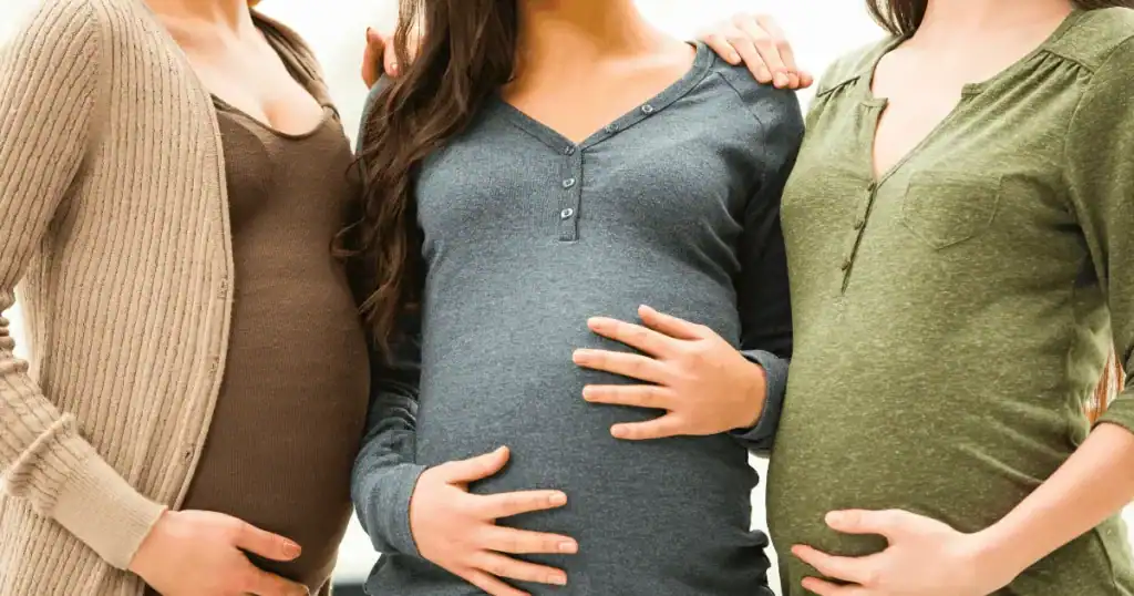 Three pregnant women standing side by side, each touching their baby bumps, wearing different colored long sleeve tops. Only their torsos are visible, highlighting the unity of expectant mothers as they share this journey—perhaps discussing essentials like the best prenatal vitamins for a healthy pregnancy.