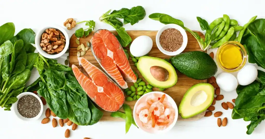 A wooden board with salmon fillets, shrimp, avocado, eggs, spinach, nuts, seeds, and edamame—rich sources of Omega 3 for brain health—surrounded by green vegetables, nuts, and a small bowl of oil on a white background; ideal for those interested in nutrition related to ADHD.