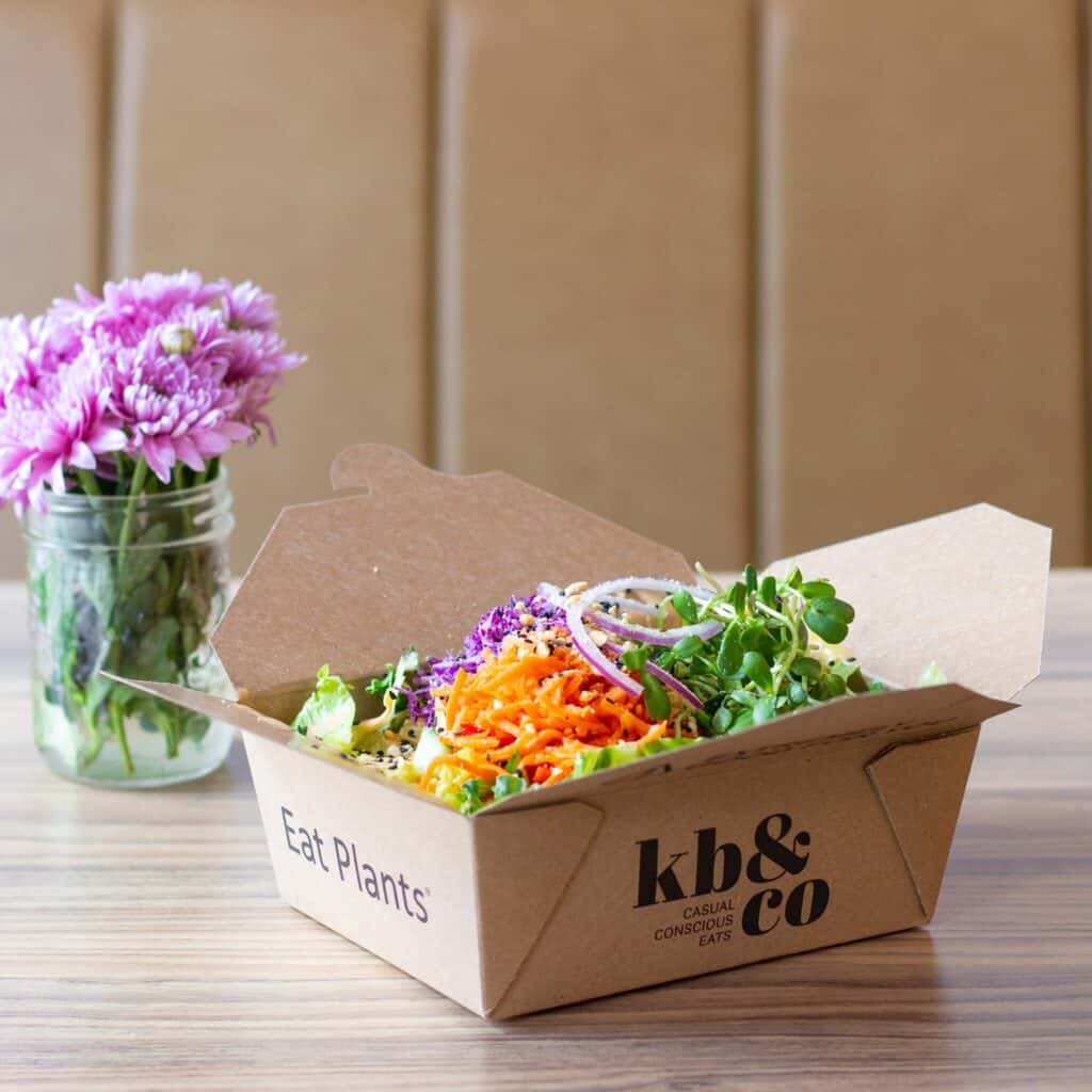A cardboard takeout box filled with a fresh salad sits on a wooden table next to a glass jar with purple flowers. The box is labeled “kb & co” and “Eat Plants.” Known as one of the top healthy restaurants in Edmonton, kb & co offers vibrant plant-based options perfect for anyone seeking nourishing meals.