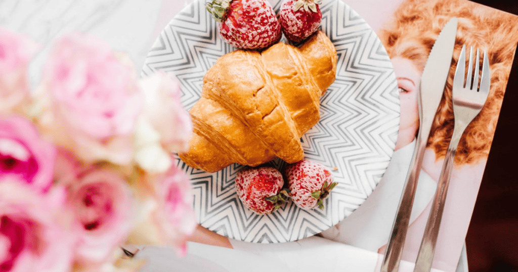 A croissant and three strawberries on a patterned plate, with a fork and knife beside it and pink flowers in the foreground—an inviting scene that gently hints at the comforting allure of emotional eating.