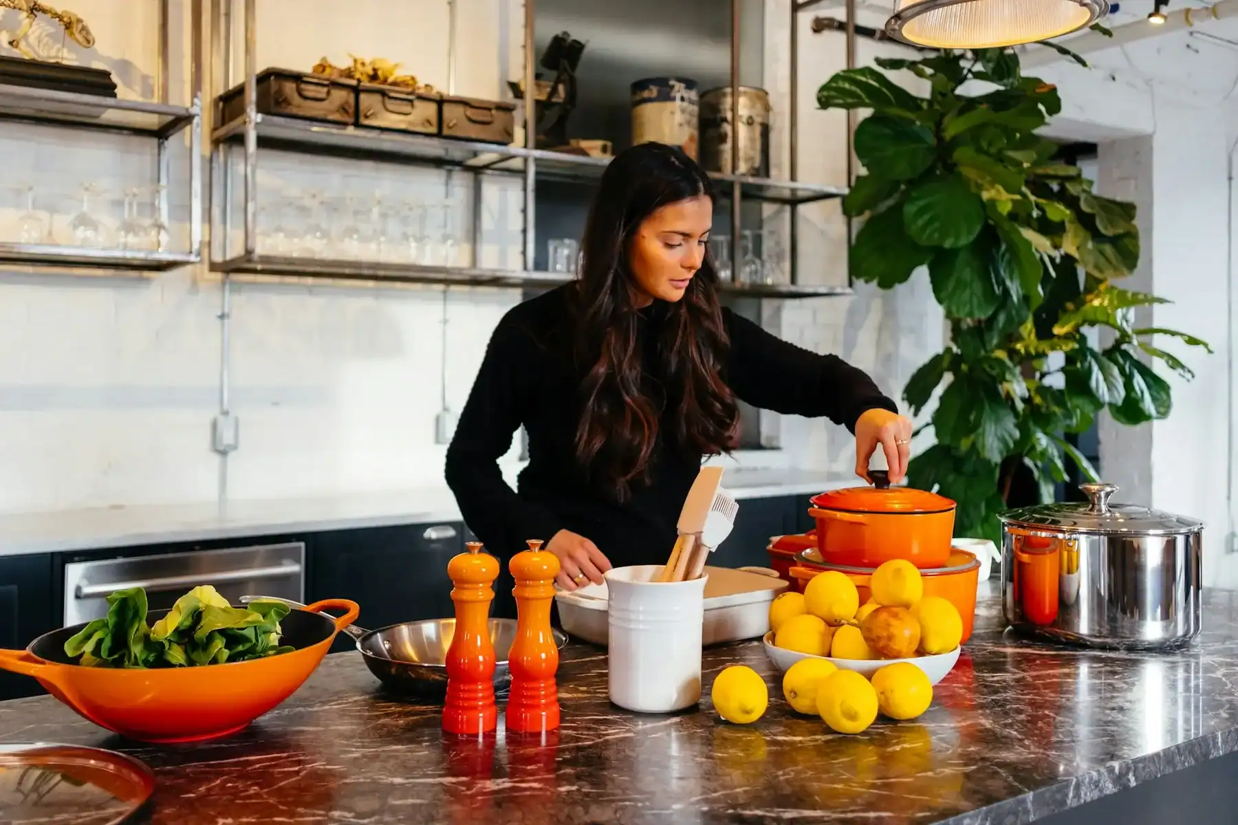 A woman in a kitchen stands at a marble counter preparing food, surrounded by lemons, spinach, kitchenware, and orange cookware. Shelves with dishes and a large plant are in the background.