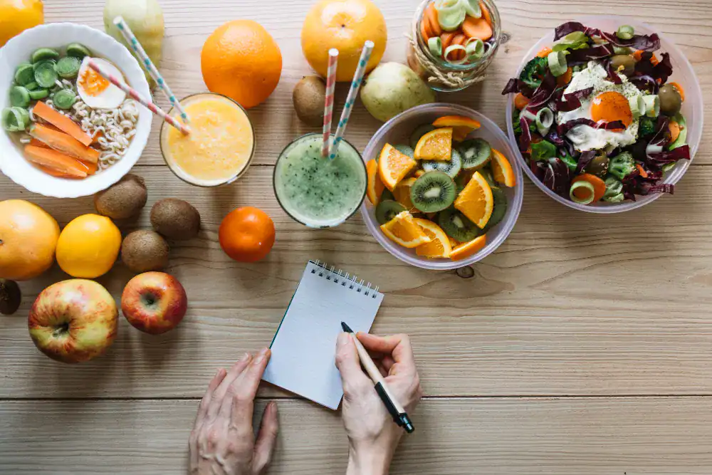 A person writes in a notepad at a table with fruit, salads, smoothies, noodles, and juice. Apples, oranges, kiwis, and greens are arranged on the wooden surface.