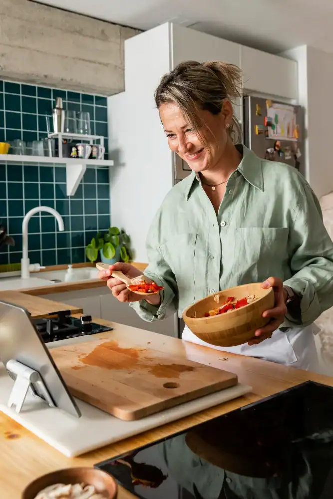 Woman in a kitchen smiles while preparing food, holding a wooden bowl and spoon, and looking at a tablet on the counter in front of her.