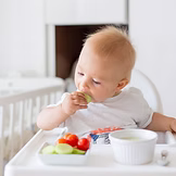 A baby sits in a high chair, eating sliced vegetables and holding food in one hand. A bowl and plate with more vegetables are on the tray in front of the baby.