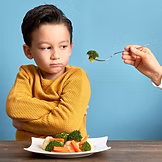 A young boy in a yellow sweater sits at a table with arms crossed, looking unhappy as an adult offers him a forkful of broccoli. A plate of vegetables is in front of him, and the background is blue.