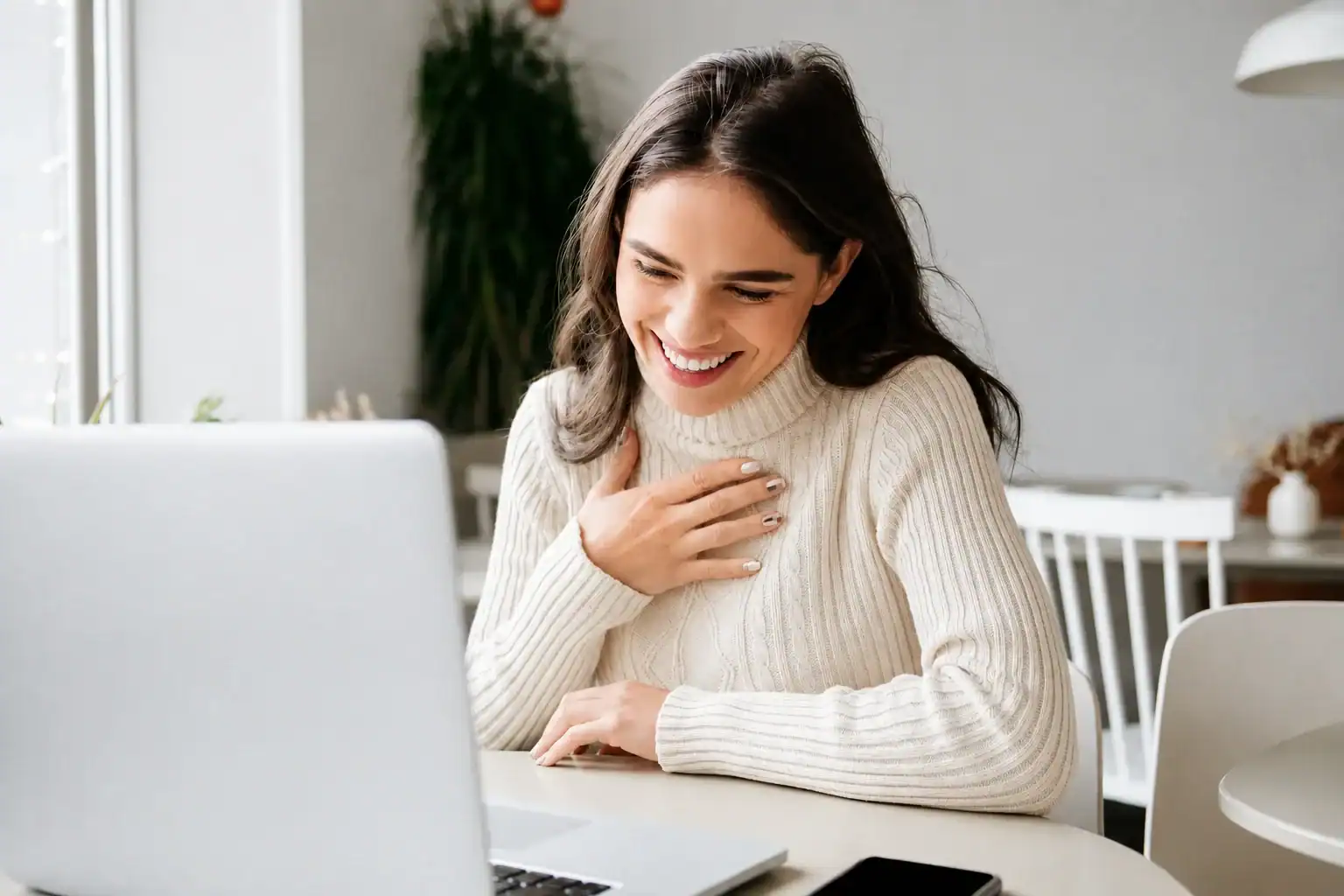 A woman in a white sweater smiles and touches her chest while looking at a laptop, sitting at a table with a smartphone, in a bright room with plants and white furniture.