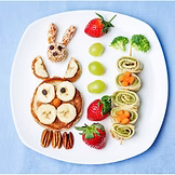 A white plate has fruit, pancakes, and rolled wraps arranged as an owl, bunny, trees, and flowers, along with strawberries, grapes, and pecans, all on a blue placemat.