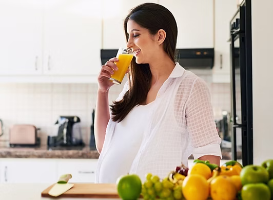 Prenatal & Postnatal Dietitians, Pregnant woman in the kitchen with vegetables on the counter smiling drinking home-made Juice