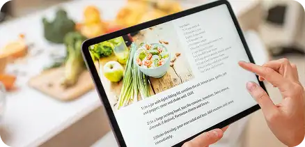 Person holding a tablet displaying a salad recipe with an image of a salad and ingredients in the background on a kitchen counter.