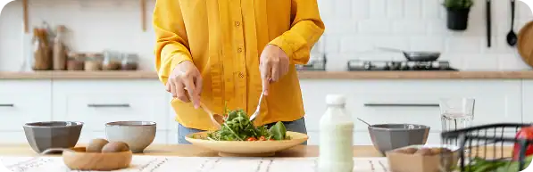 Person in a yellow shirt preparing a fresh salad in a modern kitchen, with bowls, a bottle, and fruits on the counter.