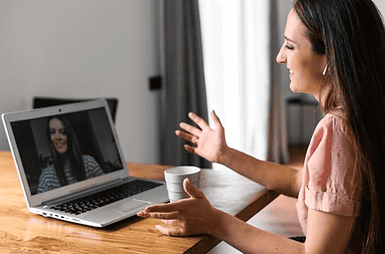 A woman sits at a table, smiling and gesturing while having a video call on her laptop. She holds a cup and wears wireless earbuds. Another woman appears on the laptop screen, smiling during the conversation.