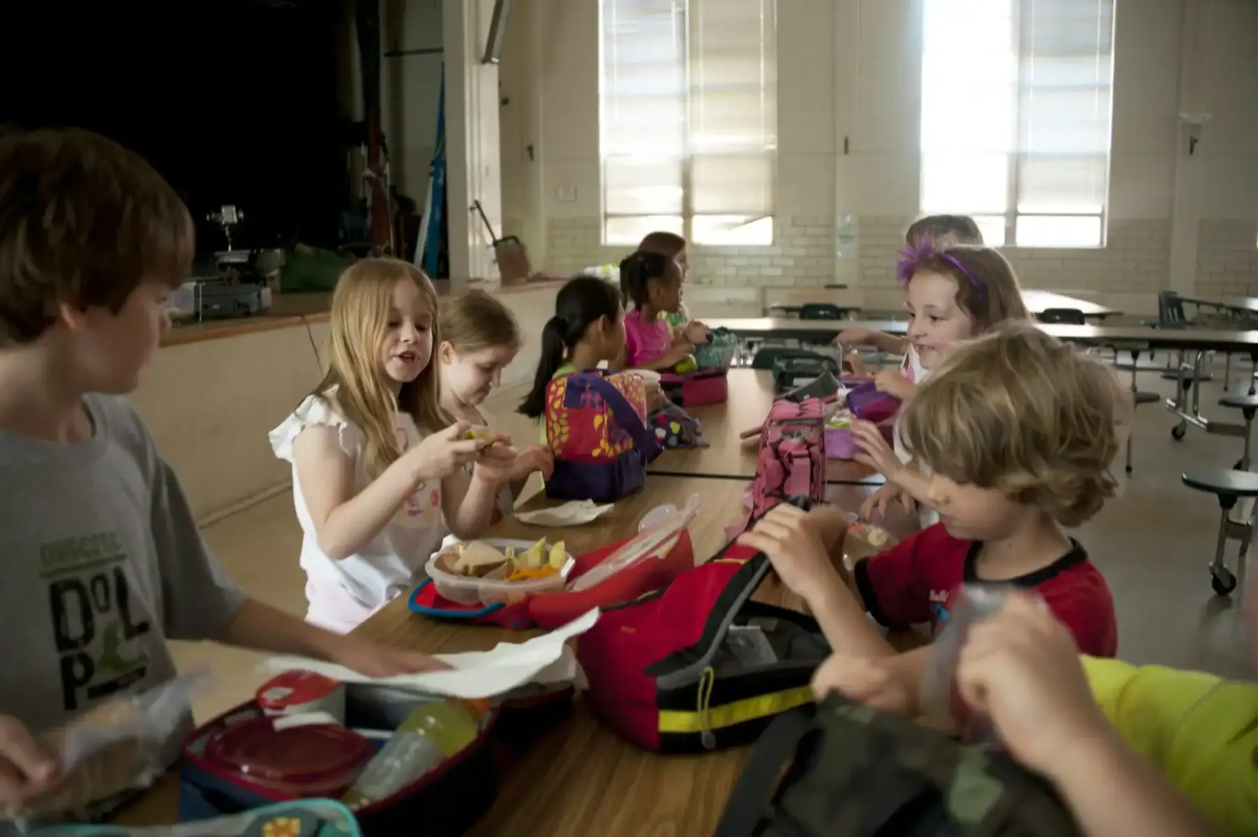 Children sit at a long cafeteria table eating lunch from lunchboxes, talking and interacting with each other in a brightly lit room with large windows.
