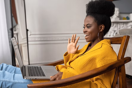 A woman in a yellow sweater sits in a chair, smiling and waving at her laptop screen during a video call.