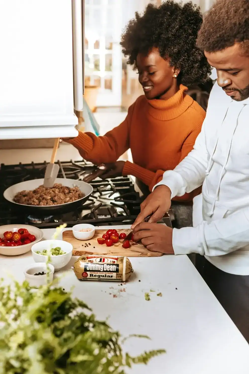 A man chops tomatoes while a woman cooks ground meat on a stove. Various vegetables and a sausage package are on the counter in a bright kitchen.