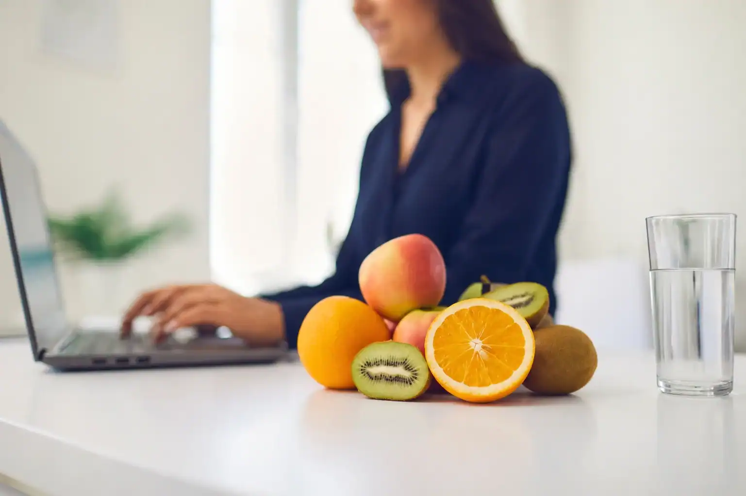 A woman works on a laptop at a desk with assorted fruits—apple, orange, kiwi—and a glass of water in the foreground.
