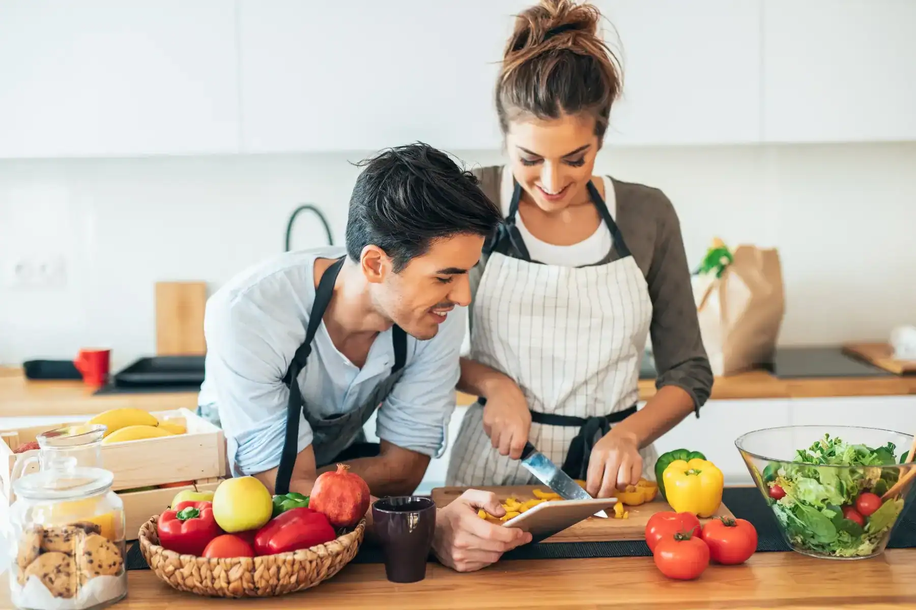 A man and woman in aprons prepare food together in a bright kitchen, chopping vegetables at a wooden counter with fresh produce and a salad bowl nearby.