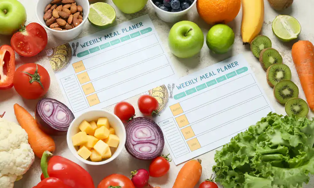 Two weekly meal planner sheets surrounded by fresh fruits and vegetables, including apples, bananas, tomatoes, carrots, lettuce, onions, kiwi, lime, almonds, blueberries, mango cubes, and cauliflower.