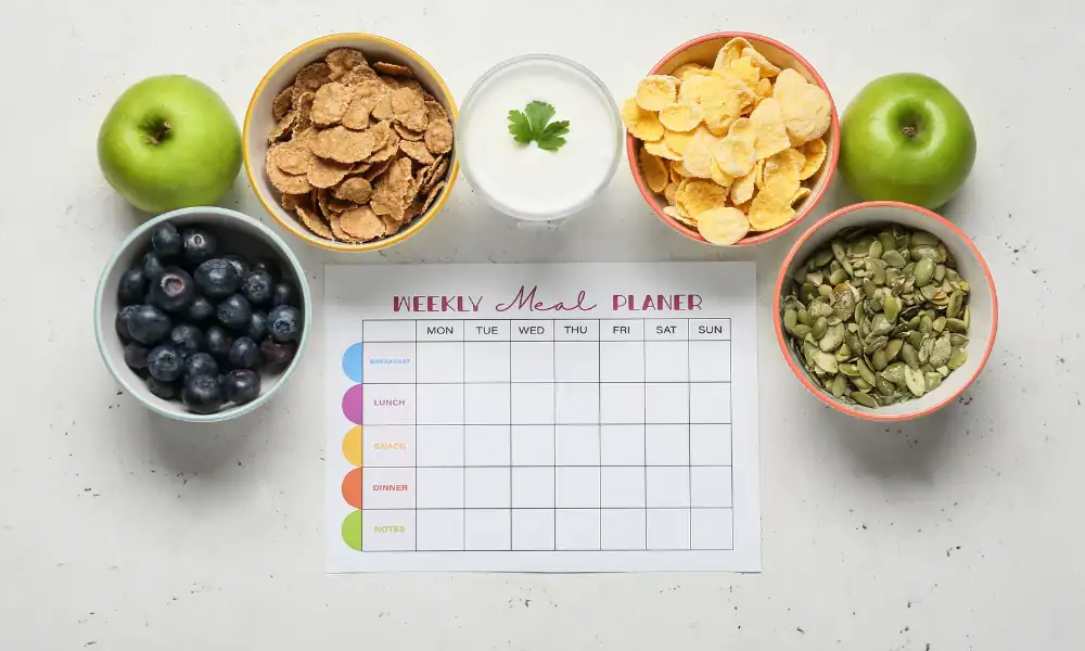 A weekly meal planner sheet is surrounded by bowls of cornflakes, bran flakes, blueberries, pumpkin seeds, a green apple, and a glass of milk on a white surface.
