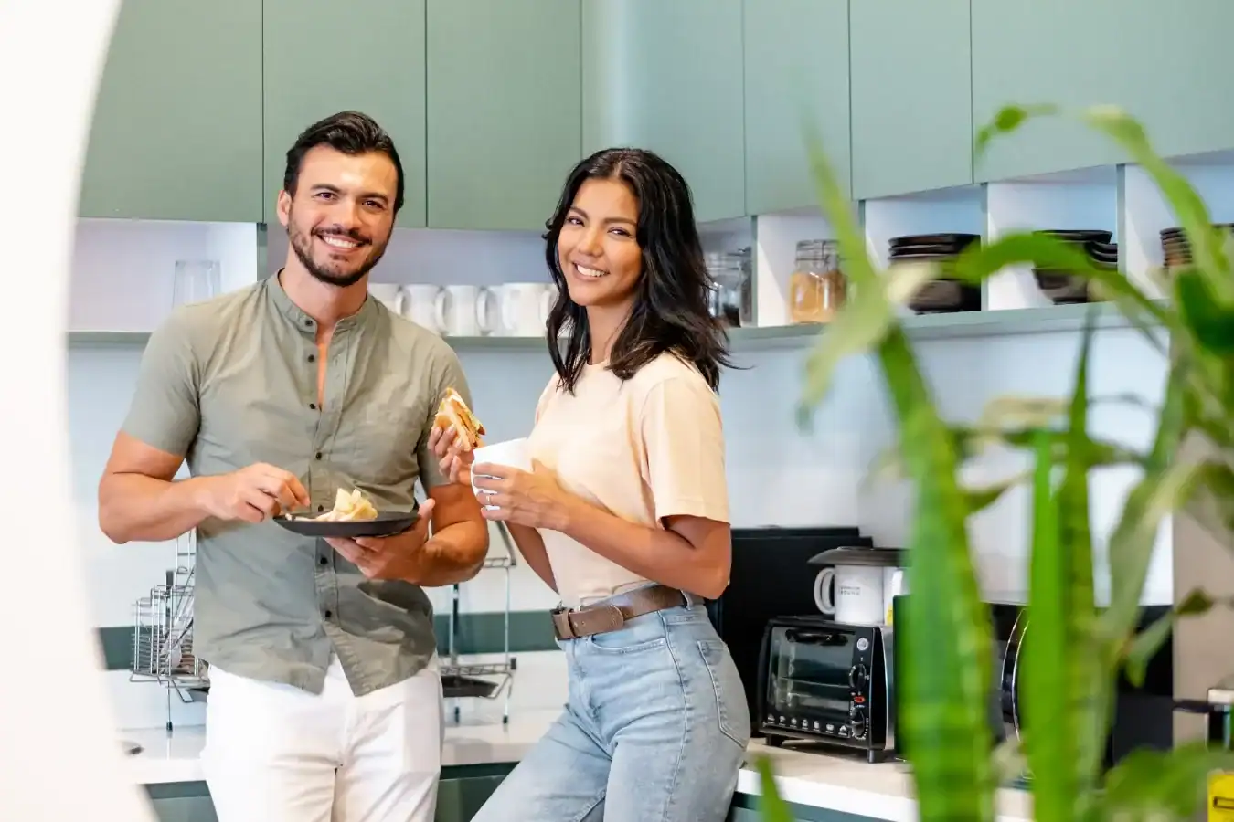 A man and woman smiling while eating pastries in a modern kitchen. The man holds a plate and fork, and the woman holds a cup. Green cabinets and kitchen appliances are visible in the background.